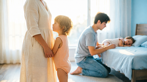 A mother and child in morning light with a parent kneeling beside a sleeping child in the background, showing the routine across generations.